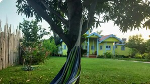 Una hamaca atada a un árbol en un patio cubierto de césped, con una casa amarilla al fondo.