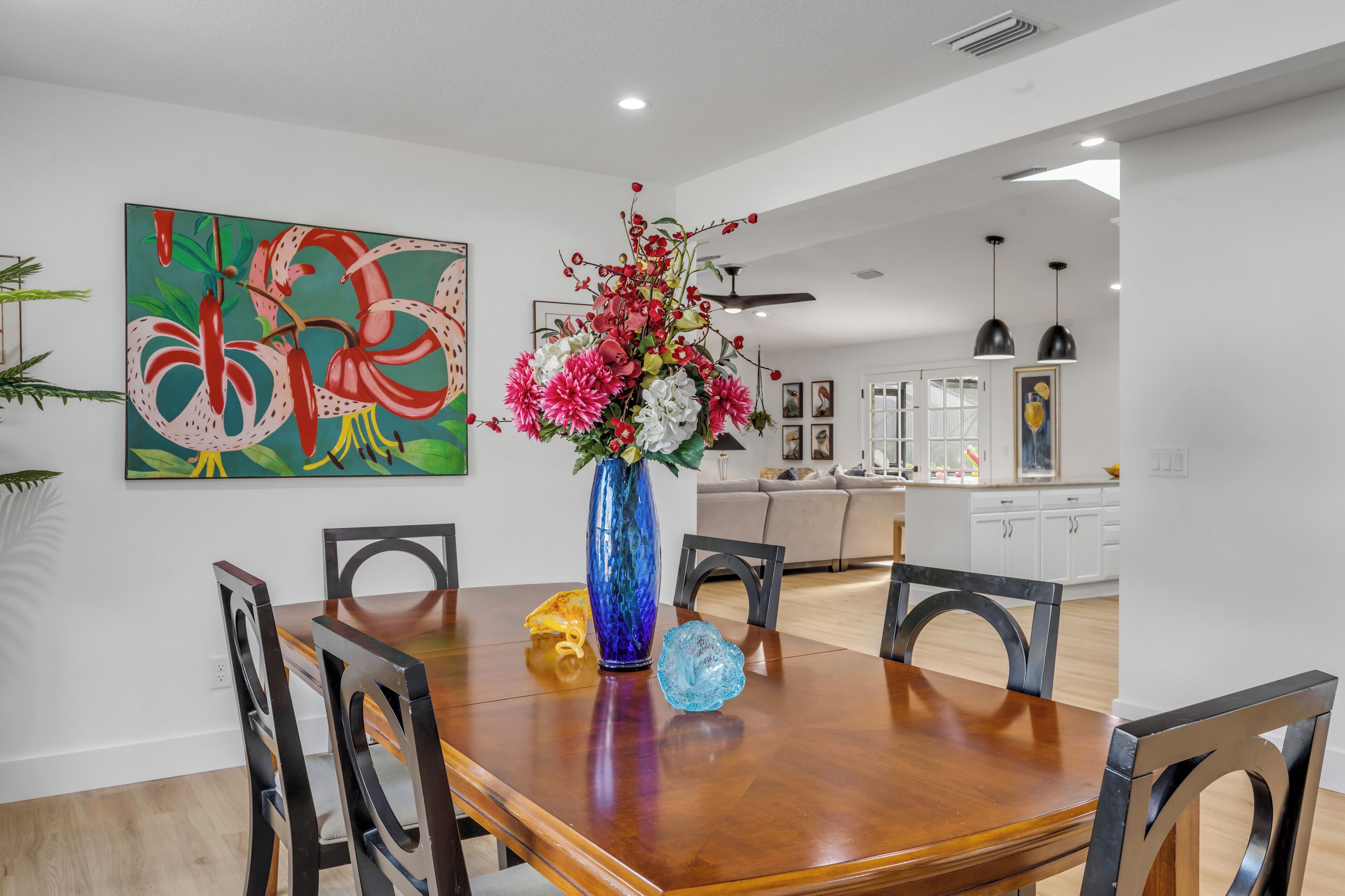 Uma sala de jantar com uma mesa de madeira, cadeiras, um vaso com flores e uma pintura abstrata colorida na parede.
