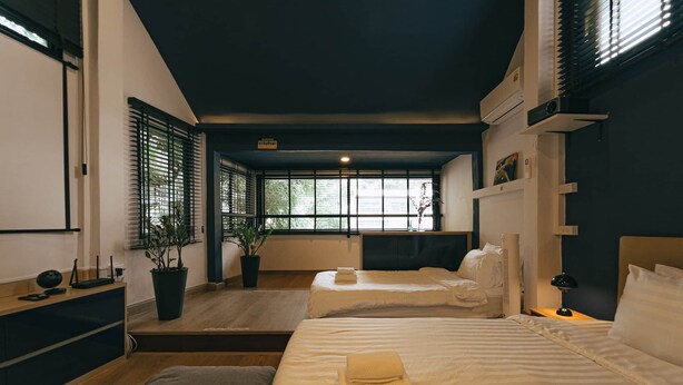 Een moderne hotelkamer met twee bedden, een groot raam met jaloezieën, een kamerplant en een nachtkastje met een lamp.