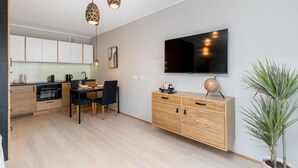 A modern kitchen with wooden cabinets, a dining area with a black table and chairs, a TV mounted on the wall, and a potted plant. - Malila Apartments Cracow Grzegórzecka 15 (Kraków)