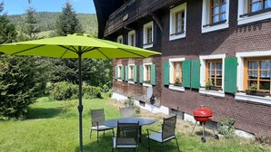 Un chalet traditionnel doté d’un patio comprenant une table et des chaises, un barbecue et un parasol vert.