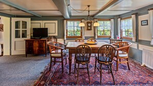 Une salle à manger avec une table en bois, des chaises, un lustre et de grandes fenêtres avec des rideaux.