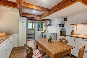 Une cuisine avec des poutres en bois, des meubles blancs, une table à manger avec des chaises et un petit îlot central agrémenté d’une plante.