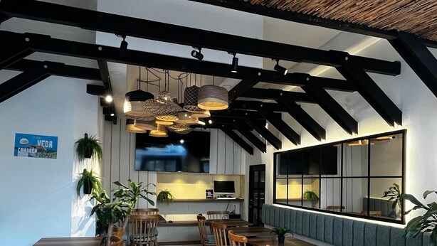 A restaurant with wooden tables and chairs, a high ceiling with exposed beams, and a kitchen area visible in the background. - Hotel La Sultana  (Tumaco)
