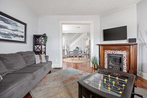 A living room with a sofa, a foosball table, a fireplace, and a TV on the wall. - Lockerbie Townhome | Steps to Mass Ave | Sleeps 8! (Indianapolis)