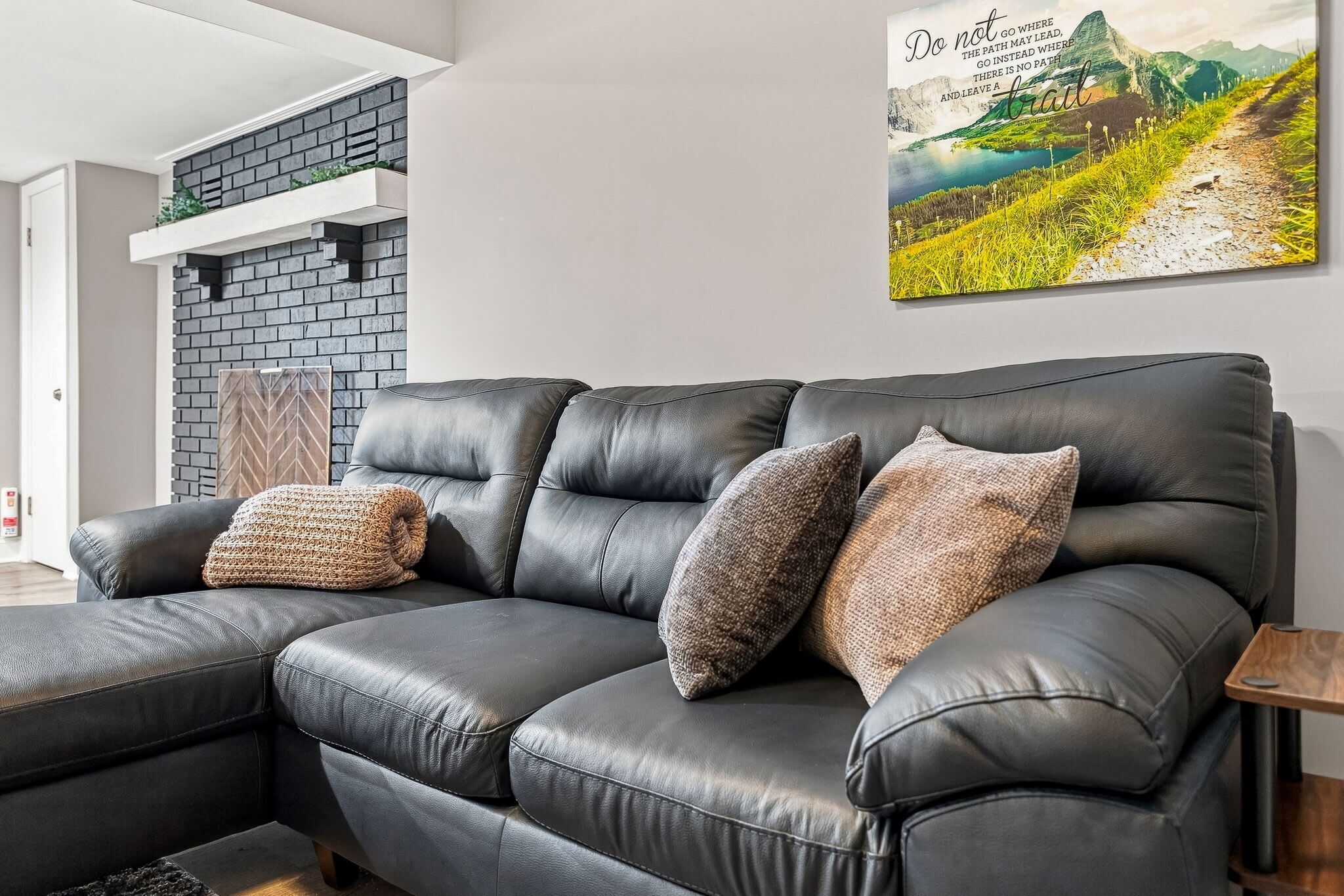 A black leather sofa with two textured pillows, a wooden side table, and a wall-mounted picture of a mountainous landscape.