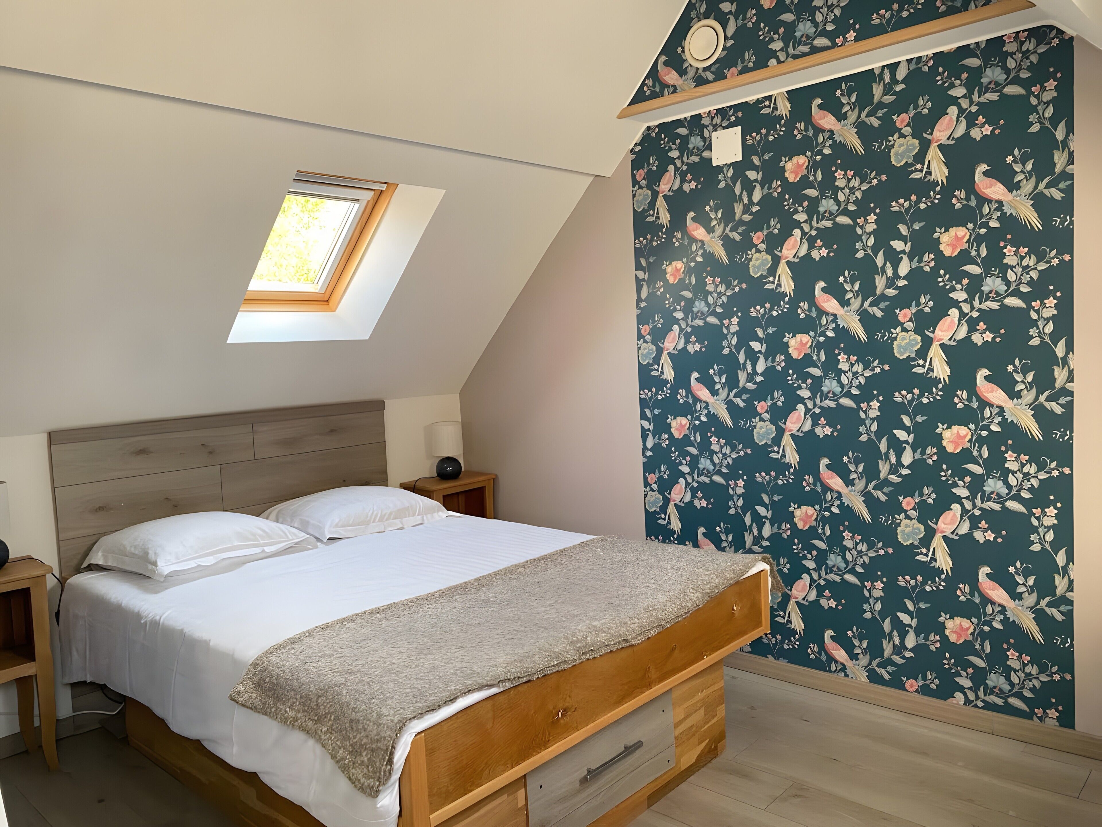 A bedroom with a skylight, a bed with a headboard, a nightstand, and a wall with a floral pattern.