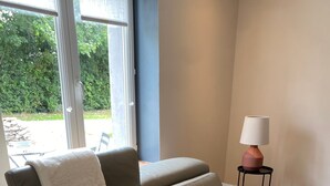 A beige leather sofa with a white throw blanket, a small side table with a lamp, and a view of greenery outside. - Holiday Home "In the Heart of Nature" with Private Terrace, Shared Garden & Wi-Fi (Ambleteuse)