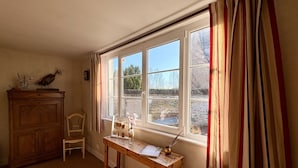 A room with a wooden desk, a chair, a window with curtains, and a peacock on a cabinet. - Charming renovated farmhouse - sleeps 6 (Boismont)