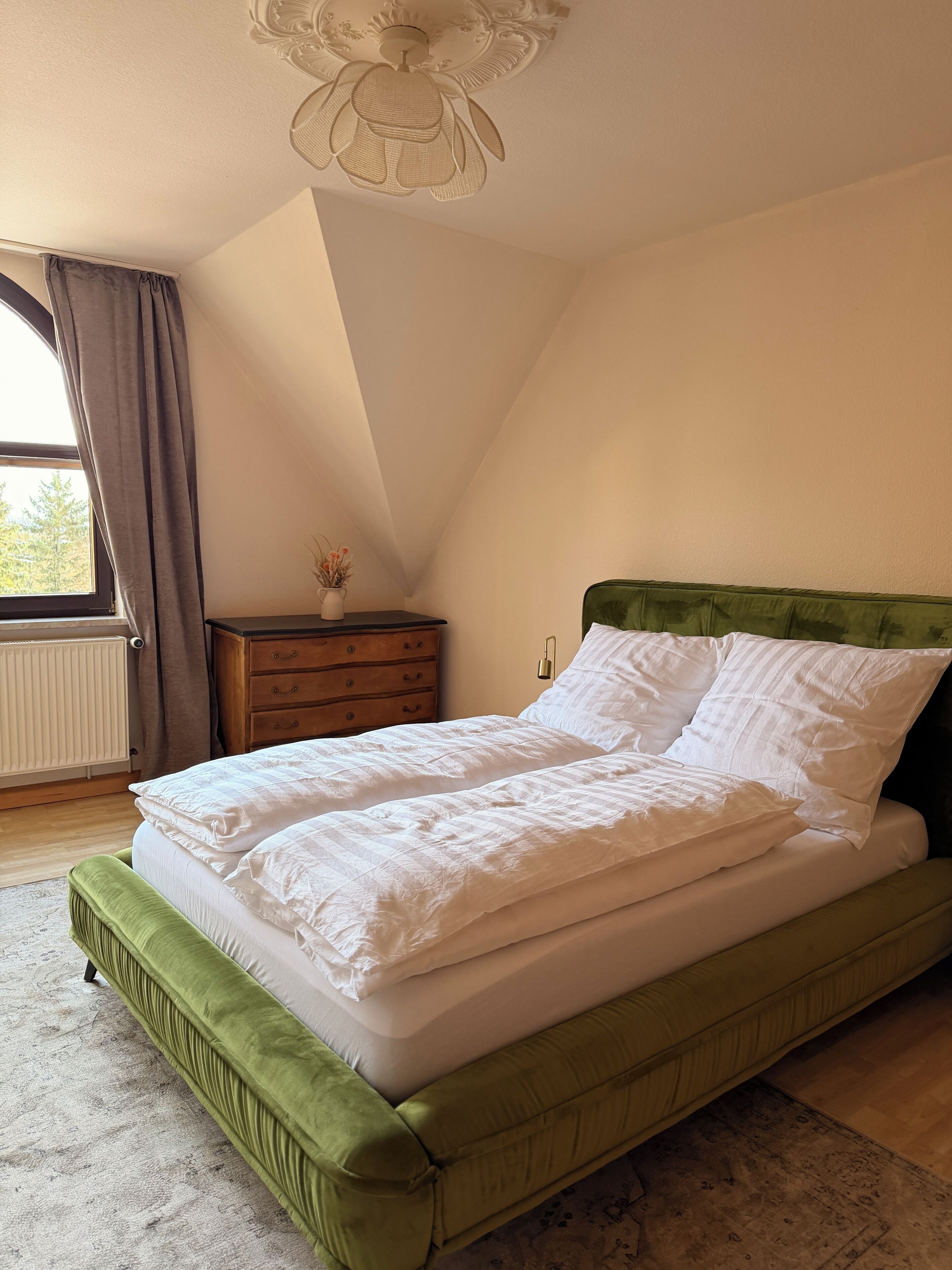A bedroom with a green upholstered bed, a wooden nightstand, a vase with flowers, a radiator, and a window with curtains.