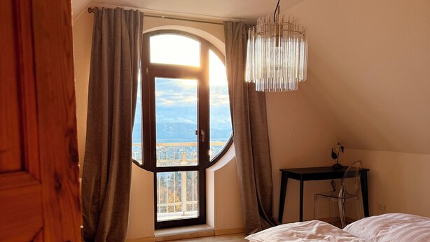 Une chambre avec une grande fenêtre, un lit, un lustre et un bureau.