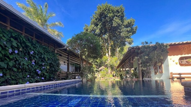 A swimming pool with blue tiles, surrounded by greenery and buildings. - Suites in a cozy house with lots of greenery and a swimming pool. (Porto Seguro)