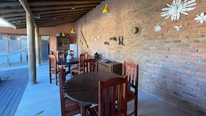 A dining area with wooden tables and chairs, a brick wall with decorative elements, and hanging yellow pendant lights. - Suites in a cozy house with lots of greenery and a swimming pool. (Porto Seguro)