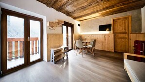 A spacious room with a wooden ceiling, large windows, a dining area with chairs, a bench, and a TV mounted on the wall. - Apt.Cristallo - `La Baita Civetta 1955` (Le Grazie)