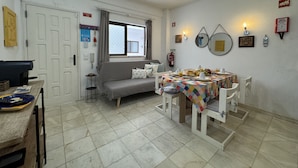 A dining area with a table set for breakfast, a sofa, and a window with curtains. - Cabanas DE Tavira Charming by Homing (Tavira)