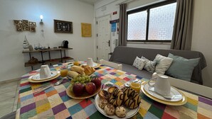 A breakfast setup with a table, breakfast items, and a couch in a room with a window and a door. - Cabanas DE Tavira Charming by Homing (Tavira)