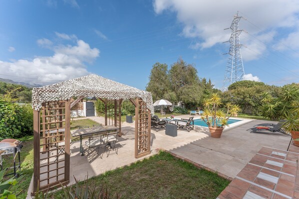 A gazebo with a lattice roof, outdoor seating, a pool, and a tall power line tower in the background.