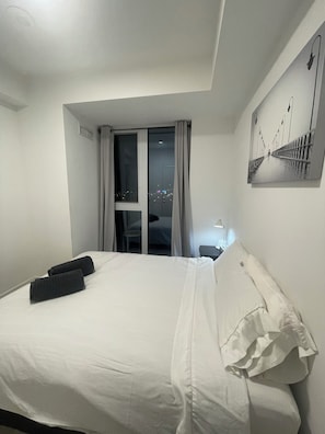 A hotel room with a bed, a window with curtains, a nightlight, and a wall-mounted artwork.
