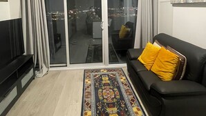A modern living room with a black sofa, a colorful rug, a TV, and a sliding glass door leading to a balcony with a city view.