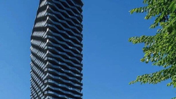A modern high-rise building with a unique zigzag facade, surrounded by a low-rise building with large windows and balconies.