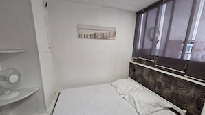 Un lit simple avec une literie blanche, une œuvre d’art accrochée au mur, une fenêtre avec des stores et une vue partielle sur une salle de bain.