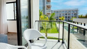 A balcony with a white wicker chair, a glass railing, and a view of a modern building and greenery. - 2 Bedrooms with Pool View (Punta Cana)