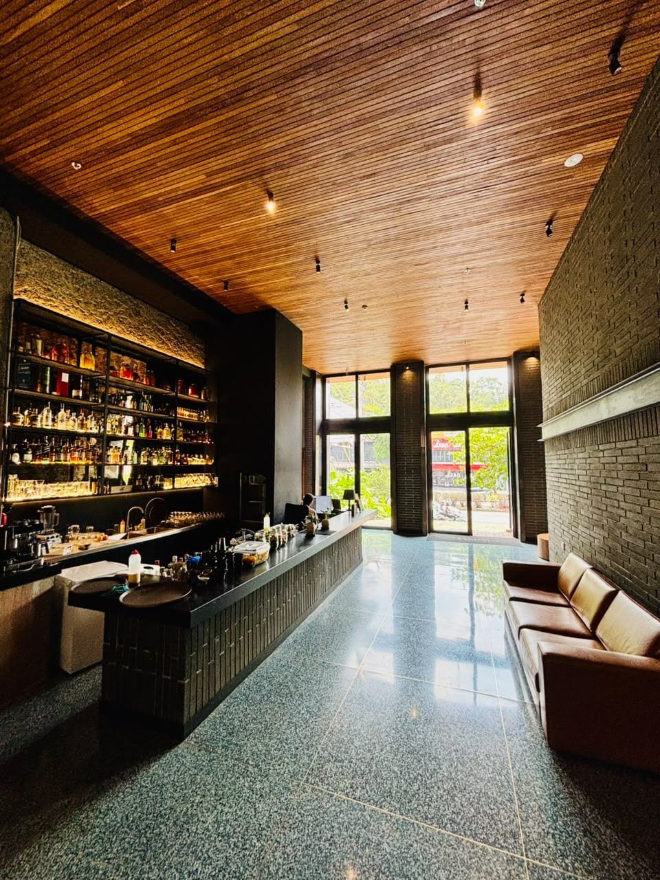 Angolo bar con soffitto in legno, bancone, scaffali con bottiglie e una zona relax con divano.