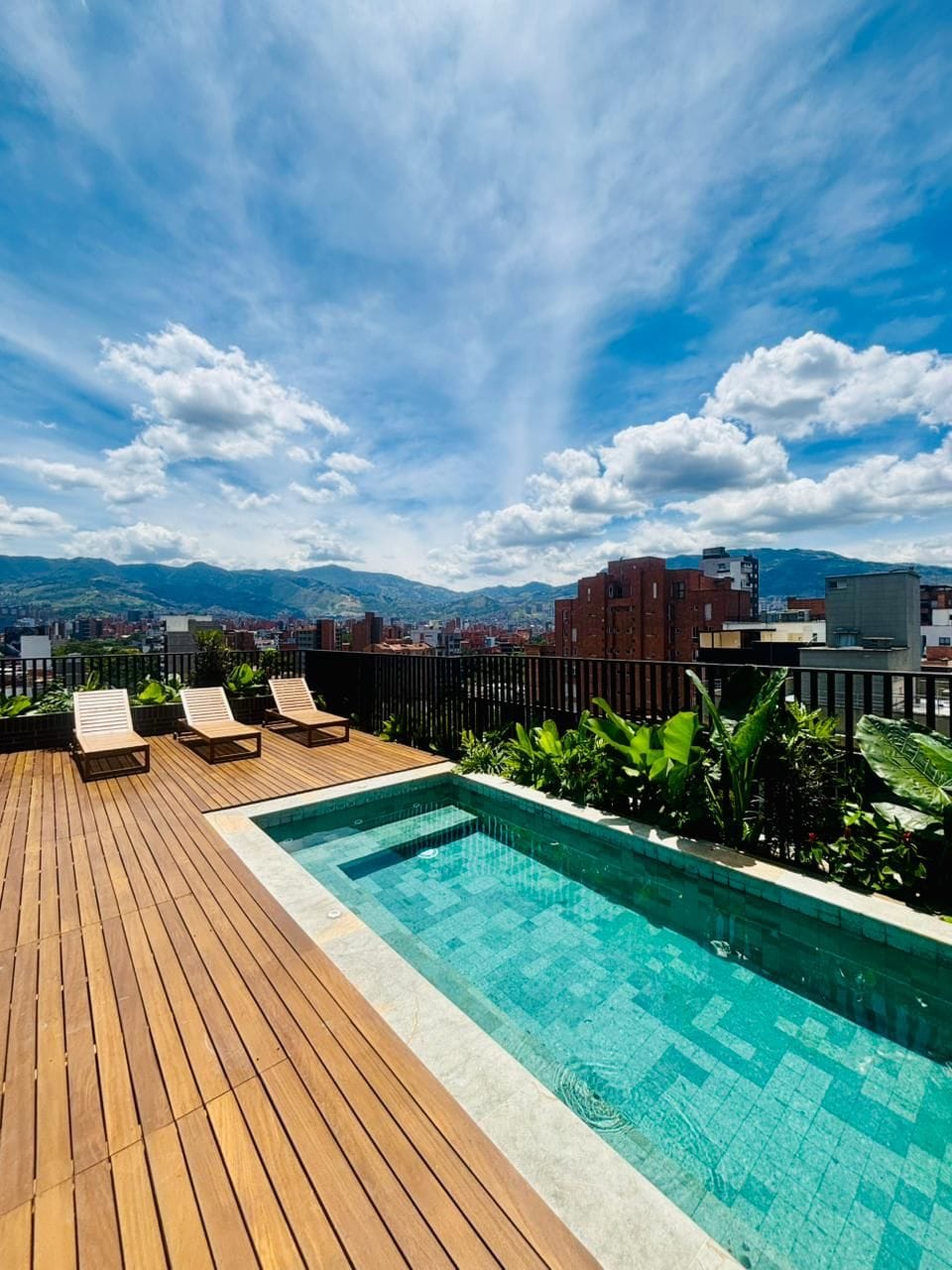 Una piscina sul tetto con terrazza in legno e poltrone relax, circondata dal verde e da un panorama urbano con montagne sullo sfondo.