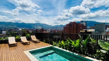 Una piscina sul tetto con terrazza in legno e poltrone relax, circondata dal verde e da un panorama urbano con montagne sullo sfondo.