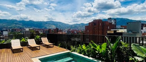 Piscina en la azotea con terraza de madera y sillas de descanso, rodeada de vegetación y un paisaje urbano con montañas al fondo.