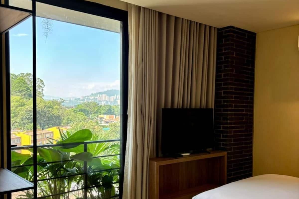 Una camera d'albergo con un letto, una televisione su un mobile in legno, un tavolo da pranzo e un'ampia finestra vista su aree verdi e edifici.