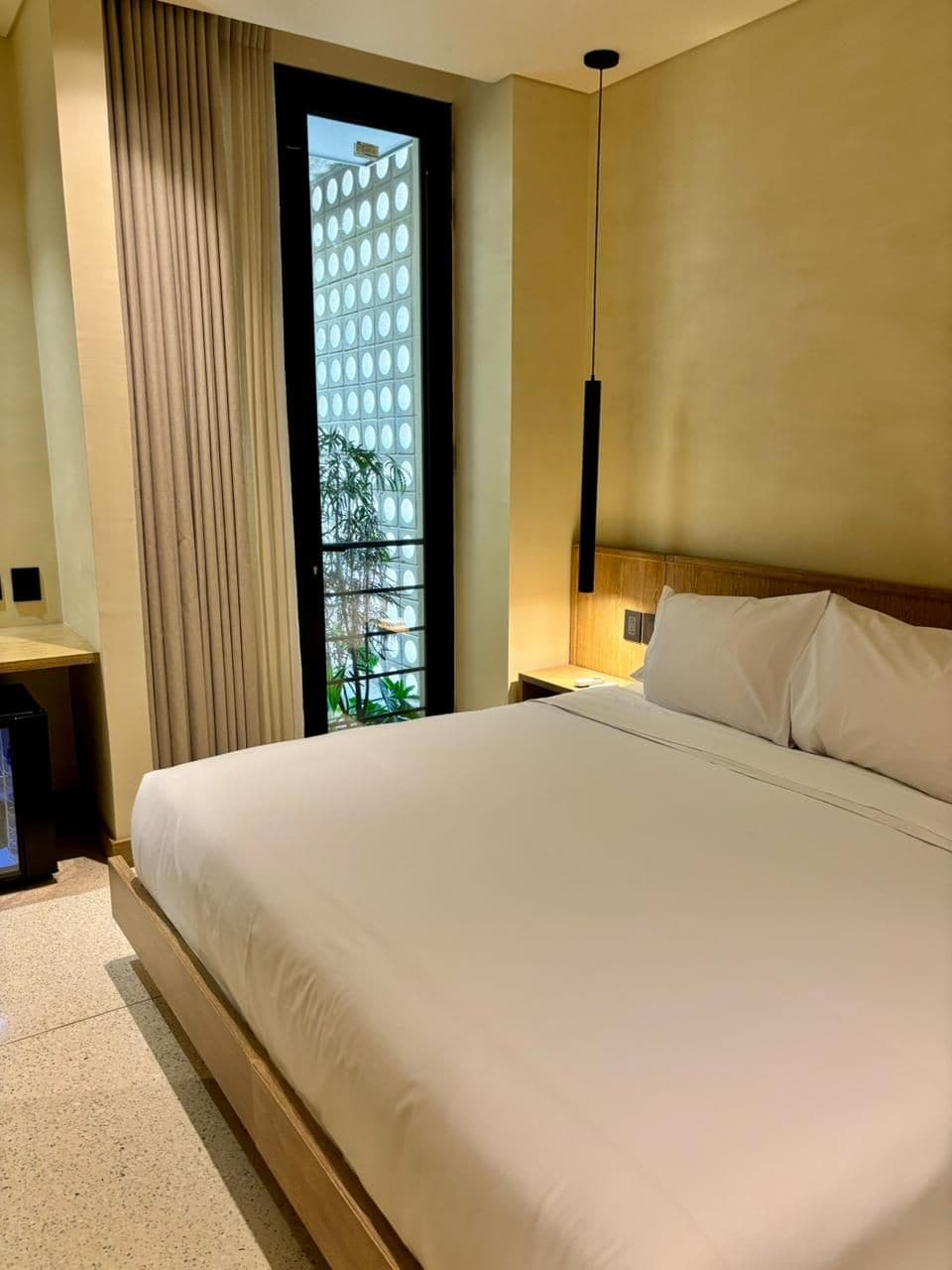 A hotel room with a large bed, a nightstand, a lamp, and a view of greenery through a window.