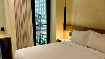 A hotel room with a large bed, a nightstand, a lamp, and a view of greenery through a window.