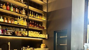 Ένας χώρος μπαρ με ράφια γεμάτα μπουκάλια με αλκοολούχα ποτά, ένας πάγκος με βάζα που περιέχουν διάφορα καρυκεύματα και μια πόρτα στο βάθος.
