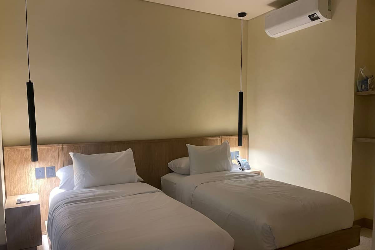 Camera d'albergo con due letti singoli, una testiera in legno, comodini con lampade e condizionatore d'aria a parete.