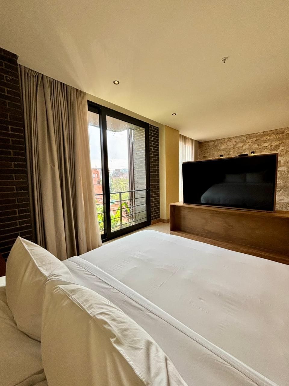 Une chambre d’hôtel avec un grand lit, un téléviseur fixé à un mur en bois, une porte-fenêtre coulissante donnant sur un balcon et un mur en briques.