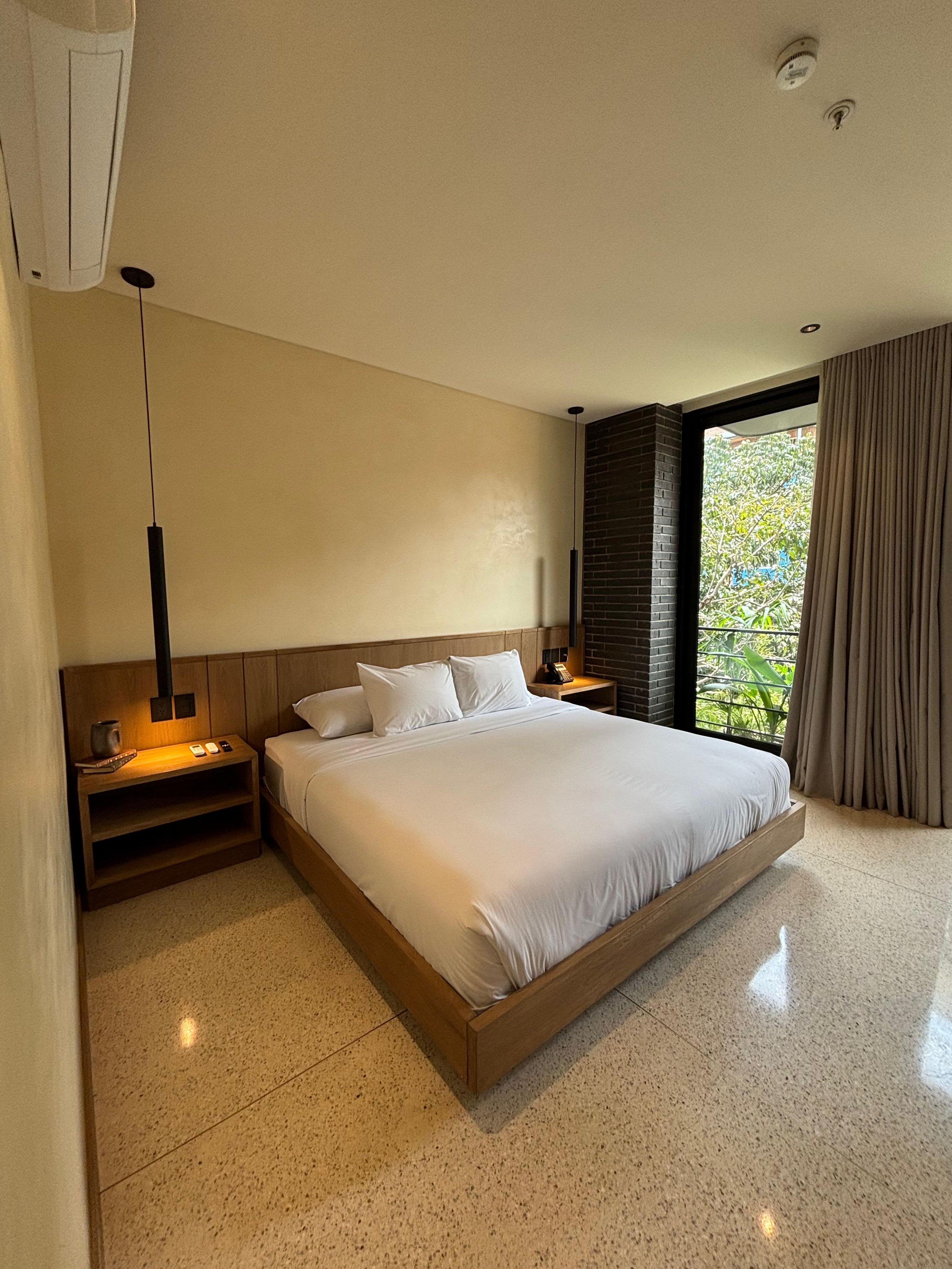 A modern bedroom with a large bed, bedside tables, a hanging light fixture, and a view of greenery through a window.