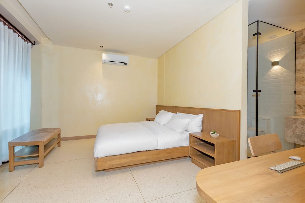 Una camera d'albergo moderna con un letto in legno, una piccola panca di legno, una scrivania con un computer e un divisorio in vetro.