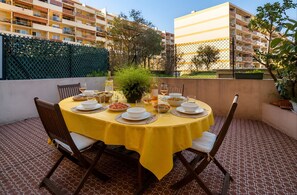 A round table with yellow tablecloth, surrounded by chairs, set on a patterned floor with a view of buildings and a green fence in the background. - Apartment "Accommodation for 4 People" with Private Terrace and Wi-Fi (Nice)