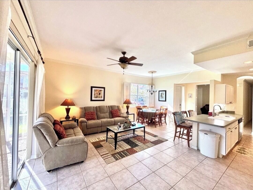 A spacious living room with a sofa, coffee table, and dining area. There is a ceiling fan and a kitchen area with a sink and cabinets.