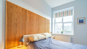 Et soveværelse med sengegavl i træ, seng med blå dyne, et natbord med lampe og et vindue med udsigt til det fri.