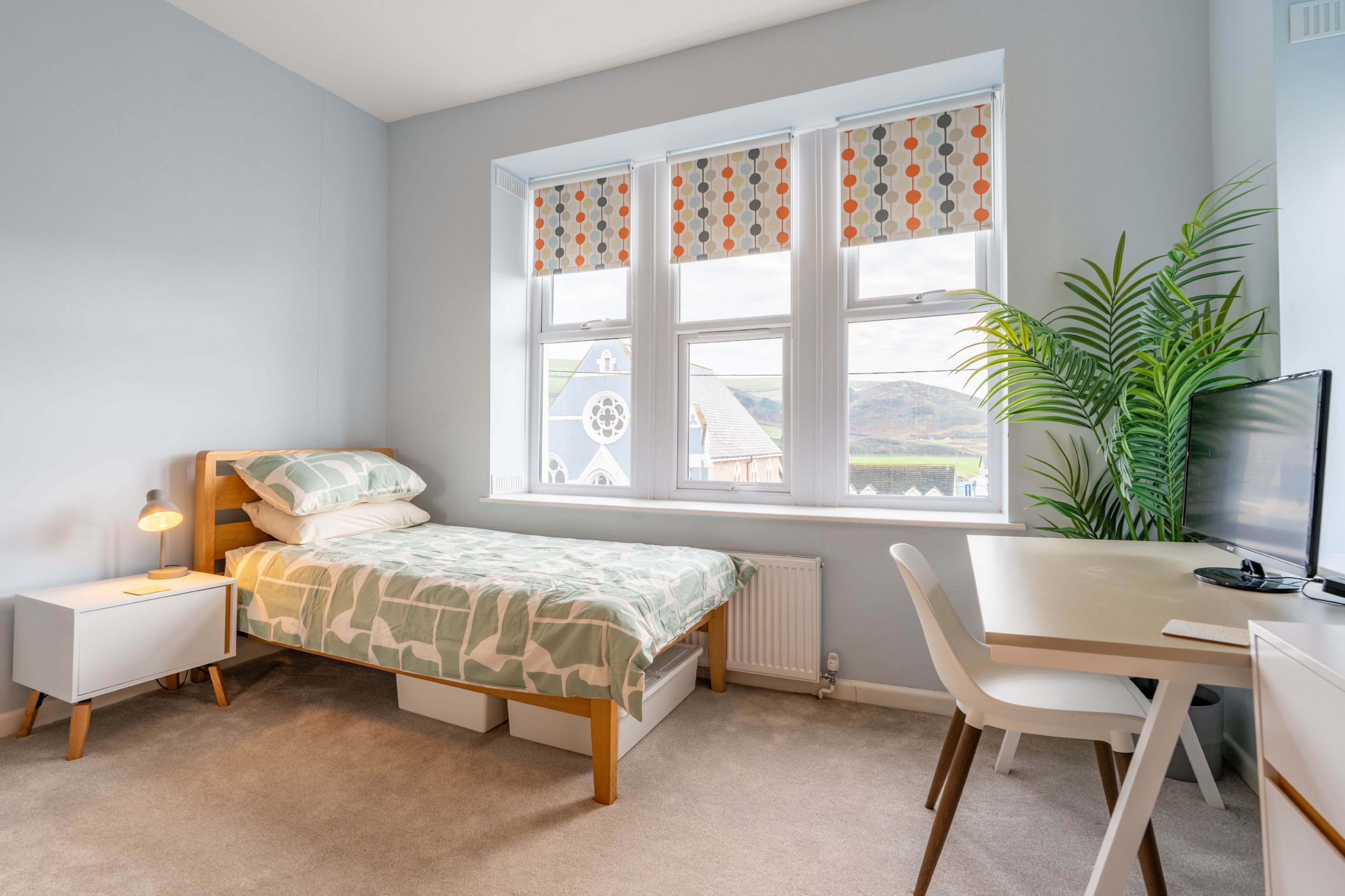 Ein Schlafzimmer mit Bett, Nachttisch, Lampe, Schreibtisch, Stuhl und einem großen Fenster mit Vorhängen.