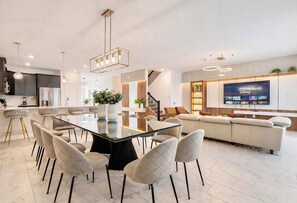 A modern kitchen with dark cabinets, a stainless steel refrigerator, and a dining area with a glass table and cushioned chairs. - ChampionsGate 9BR Retreat | Pool, Spa & Themed Rooms (Davenport)