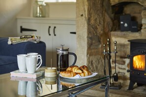 A cozy room with a glass table displaying a coffee pot, croissants, and books. A fireplace is visible in the background. - Perfect for large groups. Be it birthdays, wedding parties or a family and friends get-together, we (Bibury)