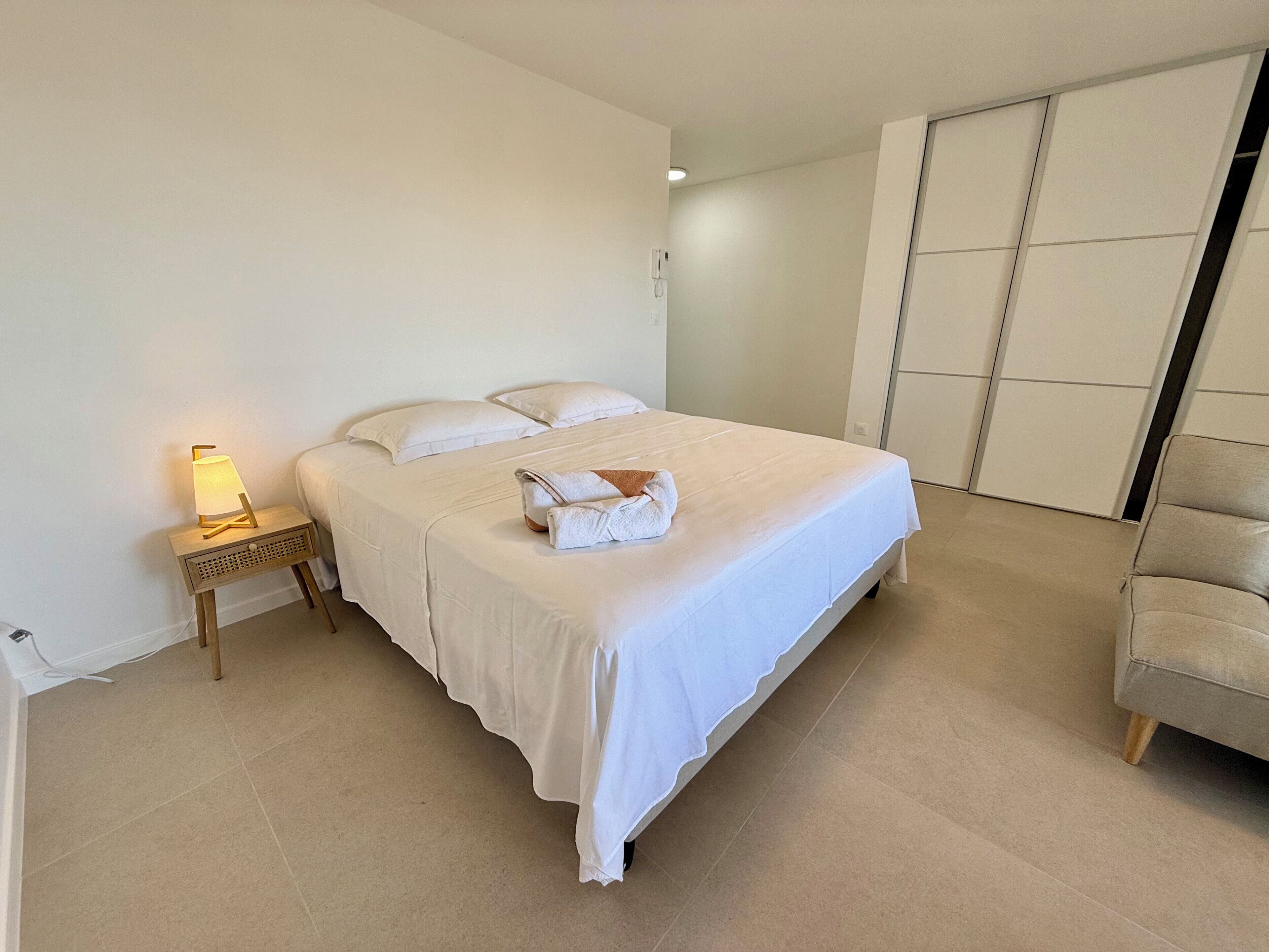 Een net opgemaakt bed met witte lakens, een bedlampje en een opgevouwen handdoek op het bed.