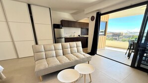 A modern living room with a sofa, two small round tables, a kitchen in the background, and a balcony with a view. - Studio Vairagi City, brand new in the center of Papeete, WiFi -Netflix -A/C (Papeete)