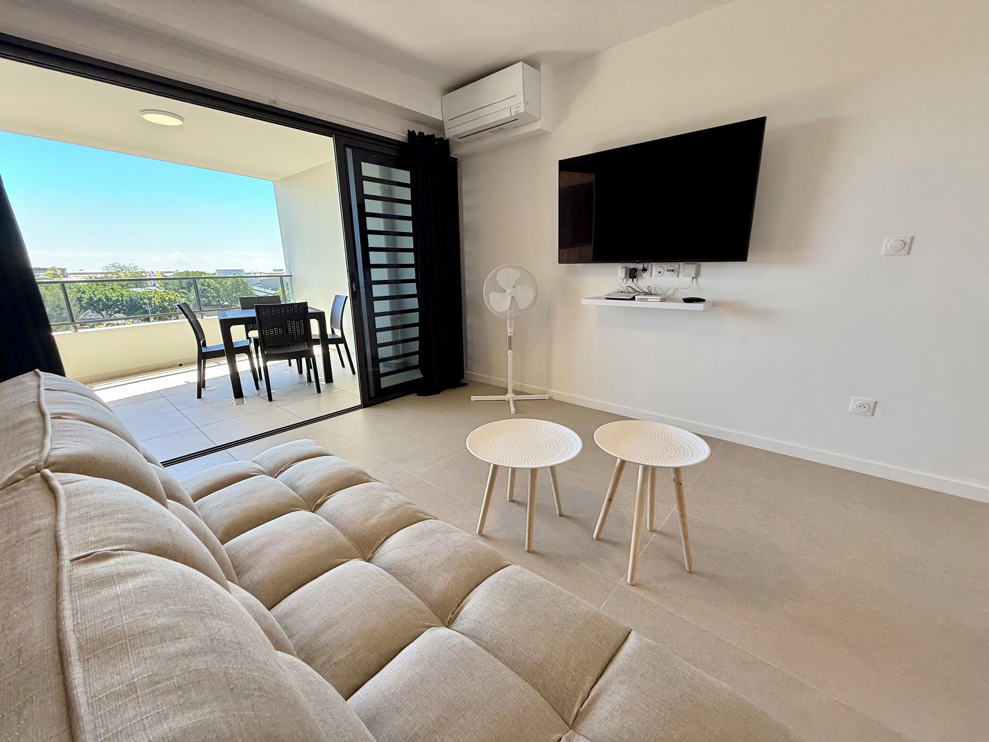 A modern living room with a sofa, two small tables, a TV, and a balcony with chairs.