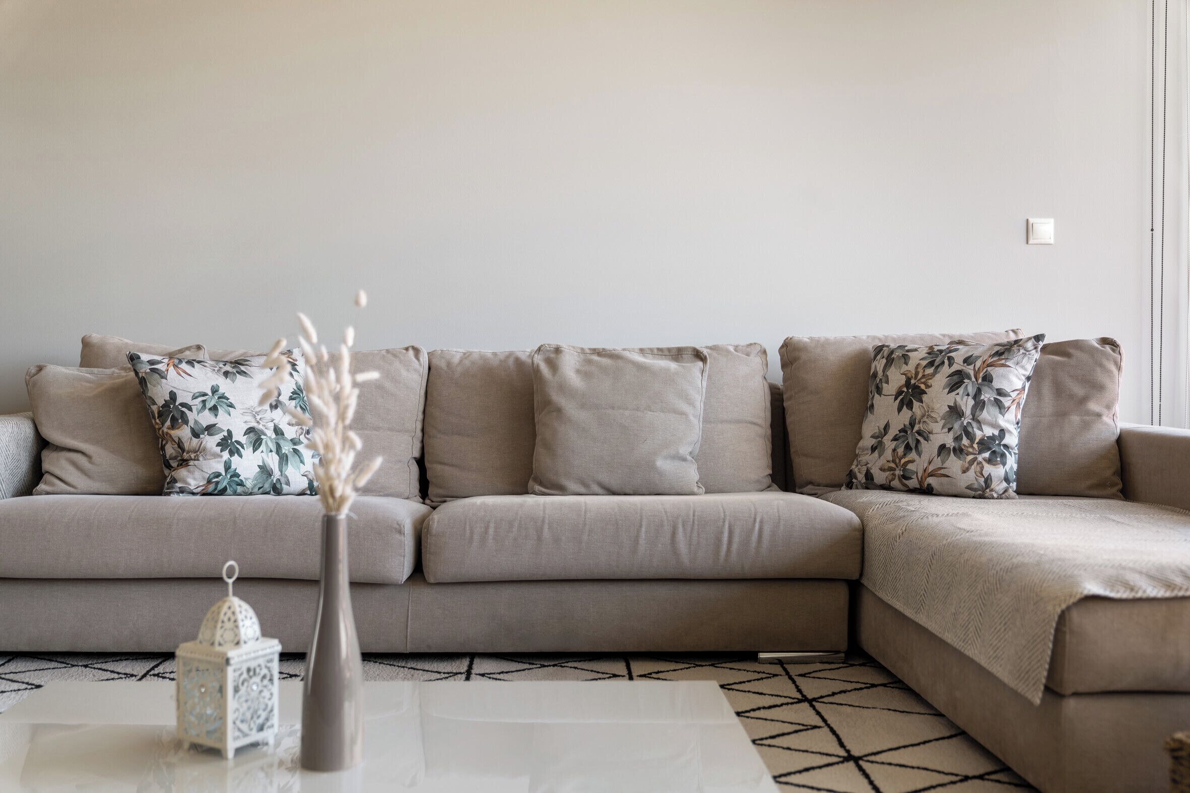 Ein beiges Ecksofa mit floralen Kissen, eine weiße Vase mit einer Dekoration und ein Teppich mit geometrischen Mustern.