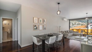 Een moderne woonkamer met een eetgedeelte, met een glazen tafel, witte stoelen, een bank en een stadsgezicht door grote ramen.