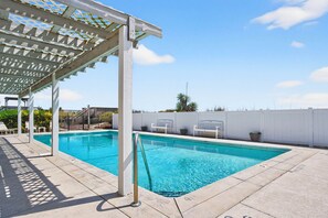 A swimming pool with a pergola, lounge chairs, and a white fence.